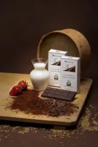 Barretta di cioccolata al latte di asina Marialuisa su una tavola di legno con confezione, giara di latte di asina fresco, polvere di cacao, fragole e setaccio in legno