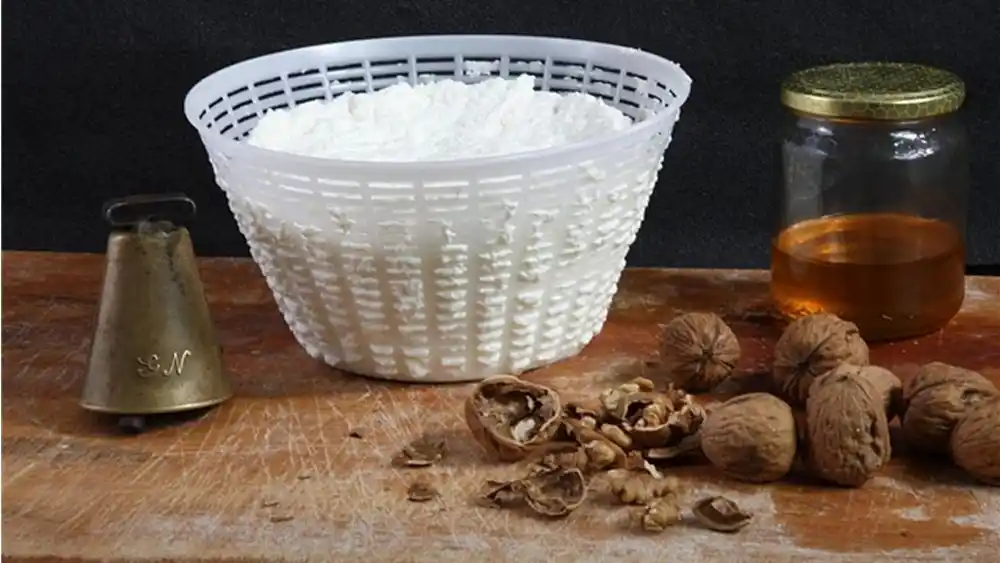 Ricotta fresca nella fuscella con miele e noci accanto a un campanello per capre su tavolo rustico